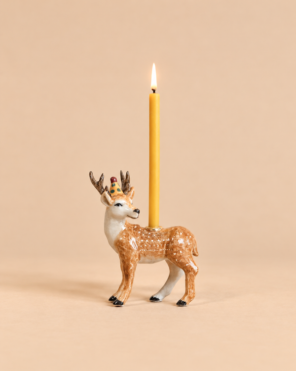 A lit yellow candle stands upright in the Stag Cake Topper, a decorative ceramic candleholder shaped like a deer with antlers and a gold crown, adorned with elegant gold accents, set against a plain beige background.