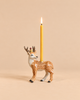 A lit yellow candle stands upright in the Stag Cake Topper, a decorative ceramic candleholder shaped like a deer with antlers and a gold crown, adorned with elegant gold accents, set against a plain beige background.