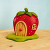 The Bumbu Handmade Wooden Strawberry House (ships in one week) is a charming red strawberry-shaped toy with a green leafy roof, wooden door, and small window, set on wood against a light blue background and inspired by Waldorf designs.