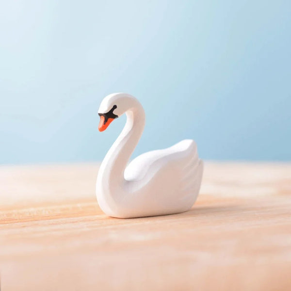 The Bumbu Handmade Wooden Swan, a small handcrafted toy with an orange beak, rests on a light wooden surface against a soft blue background.