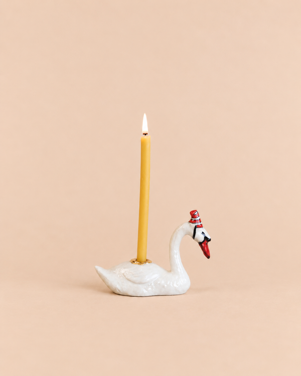 A Swan Cake Topper - Final Sale (ships in one week) features a yellow candle in a handcrafted ceramic swan holder with a red hat, glowing against a solid light peach background.