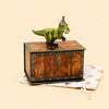A small green T-Rex Cake Topper, designed as a handcrafted collectible dinosaur figurine with a party hat, stands on a wooden treasure chest accented with gold and is displayed atop a star-patterned cloth against a beige background.