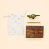 A white drawstring bag with gold stars, a T-Rex Cake Topper featuring delicate gold accents, a gold stick, and a wooden box labeled “campHollow” are arranged on a light background.