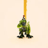 The T-Rex Cake Topper - Final Sale is a small green toy dinosaur wearing a party hat, carrying a yellow birthday candle on its back with gold accents for added whimsy, set against a plain beige background.