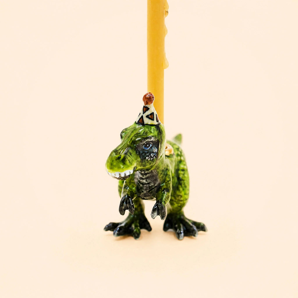 The T-Rex Cake Topper - Final Sale is a small green toy dinosaur wearing a party hat, carrying a yellow birthday candle on its back with gold accents for added whimsy, set against a plain beige background.