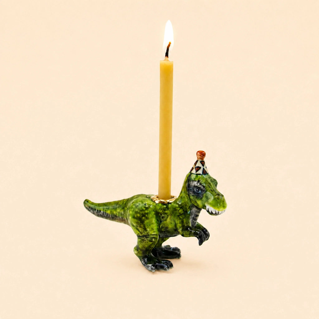 A delightful handcrafted collectible, the T-Rex Cake Topper - Final Sale features a small green dinosaur figurine sporting a party hat and carrying a lit candle on its back against a simple beige background, making it an enchanting porcelain cake topper.