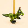 The T-Rex Cake Topper - Final Sale features a green dinosaur figurine with a party hat and a candle on its back, highlighted by gold accents and displayed against a simple light background. This delightful handcrafted porcelain piece makes for a unique collectible or cake topper.