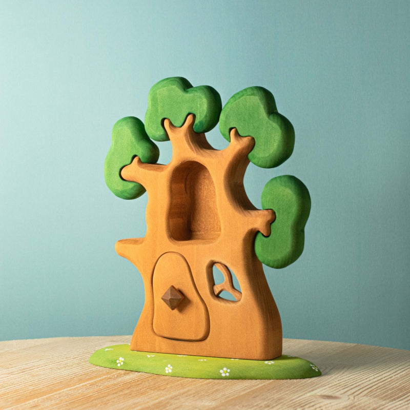 The Bumbu Handmade Wooden Ancient Oak Tree with Owl Set features green foliage, a hollow center, door, and heart-shaped window on a flat base—an inspiring playset displayed on wood with a light blue background.