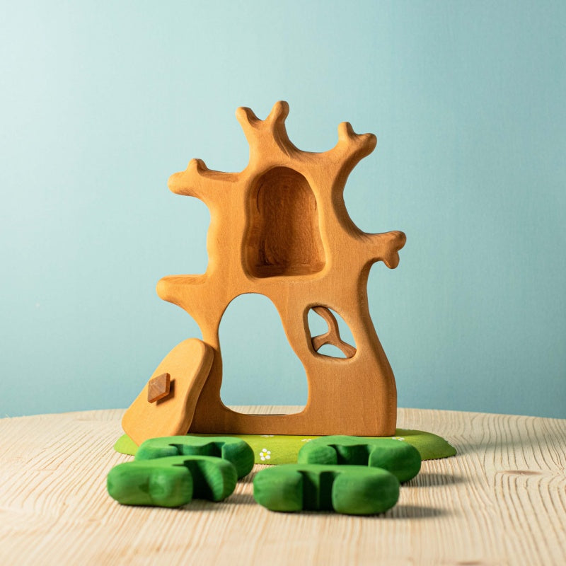 The Bumbu Handmade Wooden Ancient Oak Tree with Owl Set features a carved opening, small door, green base, and separate bush pieces for imaginative play. This sustainable maple wood toy is shown on a wooden surface with a light blue background.