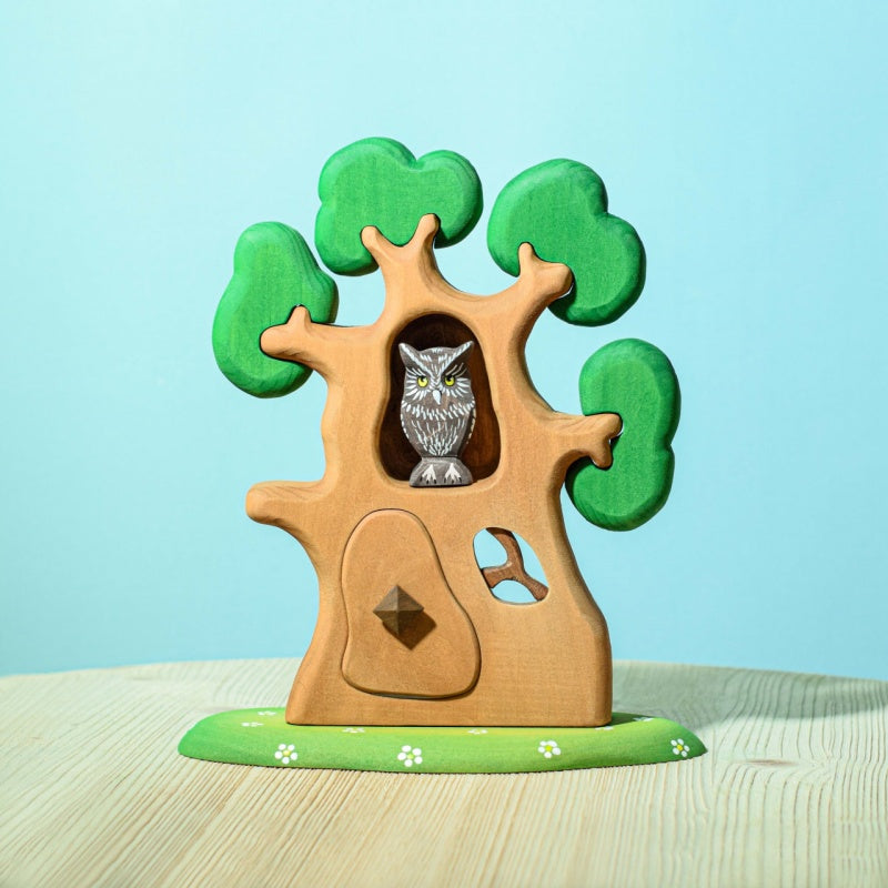 The Bumbu Handmade Wooden Ancient Oak Tree with Owl Set is a handcrafted wooden toy tree with green leaves, an owl in a hollow, white flowers on a green base, and a blue background.
