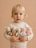 A young child with blonde hair in pigtails holds five Little Forest Animals — The Bellevue Family dolls—soft plush bunnies dressed in different outfits made from organic cotton—against a beige background.