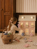 A young girl sits on the floor, playing with a pink dollhouse. She carefully arranges Little Forest Animals — The Bellevue Family handcrafted dolls and bunny dolls in front of it. A rattan gift basket filled with organic cotton dolls and doll clothes is nearby. Warm light glows from the dollhouse windows.