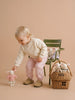 A young child in light clothes picks up a member of the Little Forest Animals — The Bellevue Family. Nearby, a green chair and a rattan gift basket hold more soft friends from the collection, all set against a beige background.