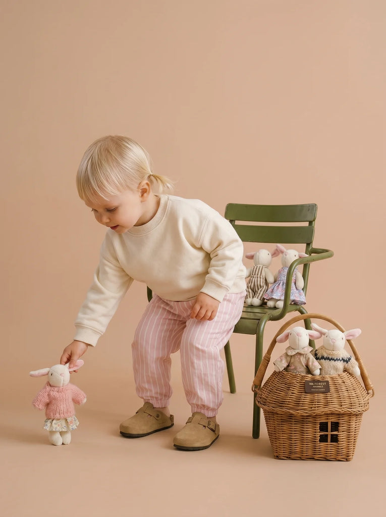 A young child in light clothes picks up a member of the Little Forest Animals — The Bellevue Family. Nearby, a green chair and a rattan gift basket hold more soft friends from the collection, all set against a beige background.