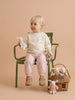 A young child with blonde hair sits on a green chair, holding an organic cotton bunny doll. The child wears a cream sweater, pink striped pants, and tan shoes. Nearby on the floor rests a rattan gift basket filled with handcrafted Little Forest Animals — The Bellevue Family dolls.