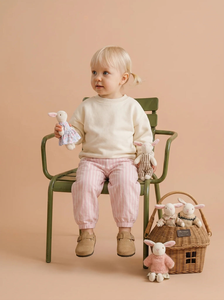 A young child with blonde hair sits on a green chair, holding an organic cotton bunny doll. The child wears a cream sweater, pink striped pants, and tan shoes. Nearby on the floor rests a rattan gift basket filled with handcrafted Little Forest Animals — The Bellevue Family dolls.