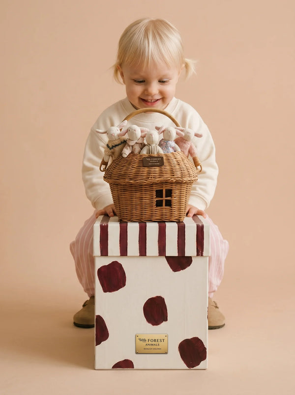 A smiling toddler with blonde hair holds the Little Forest Animals — The Bellevue Family rattan gift basket of stuffed animals while sitting on a white box with maroon spots, set against a beige background.