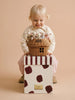 A smiling toddler with blonde hair holds the Little Forest Animals — The Bellevue Family rattan gift basket of stuffed animals while sitting on a white box with maroon spots, set against a beige background.