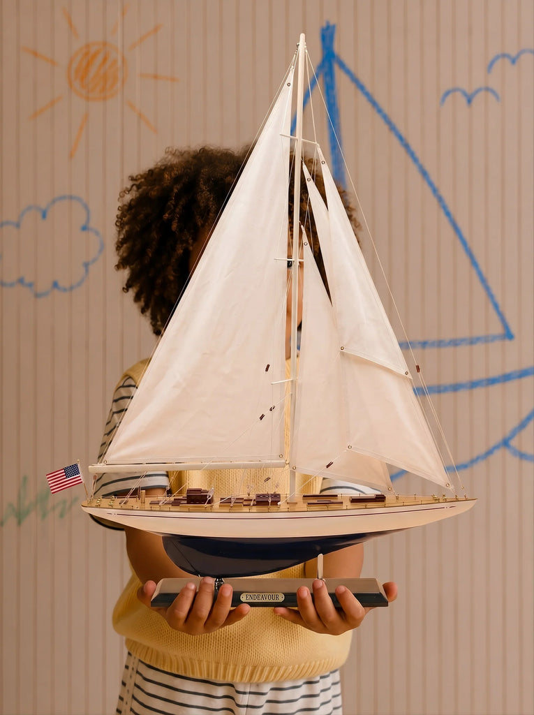A child holding "Little Forest Animals Silk Rug — The Regatta Series — The Endeavour" with white sails obscures their own face behind the model. On the beige wall behind them, there are hand-drawn illustrations of a sun, a cloud, and a blue sailboat.