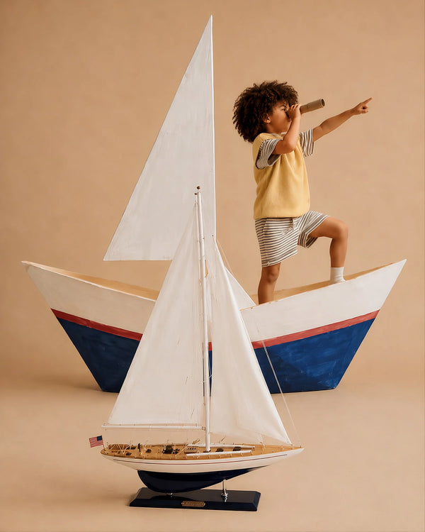 A child in a yellow vest and striped shorts stands in a cardboard sailboat, holding a telescope and pointing ahead, with the Little Forest Animals Silk Rug — The Regatta Series — The Endeavour displayed in the foreground against a beige background.