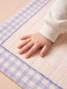 A small child's hand rests on the Little Forest Animals Silk Rug — The Gingham, a soft, hand-tufted rug featuring pastel-colored purple and white gingham borders with light pink stripes.
