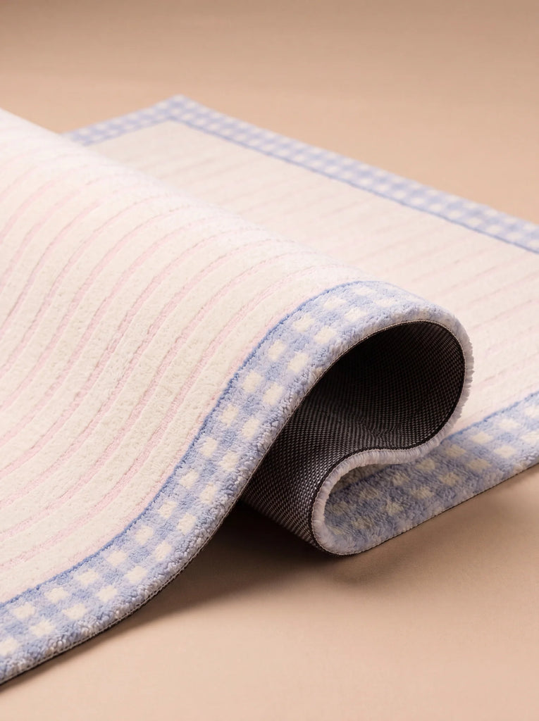 A close-up of the Little Forest Animals Silk Rug — The Gingham, a rectangular hand-tufted rug featuring a pink striped pattern with a blue gingham border and a non-slip black backing, partially rolled over a beige surface.