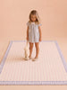 A young girl in a light blue dress stands on the Little Forest Animals Silk Rug — The Gingham, which features a charming blue gingham border, while holding a plush bunny. The background is plain beige.