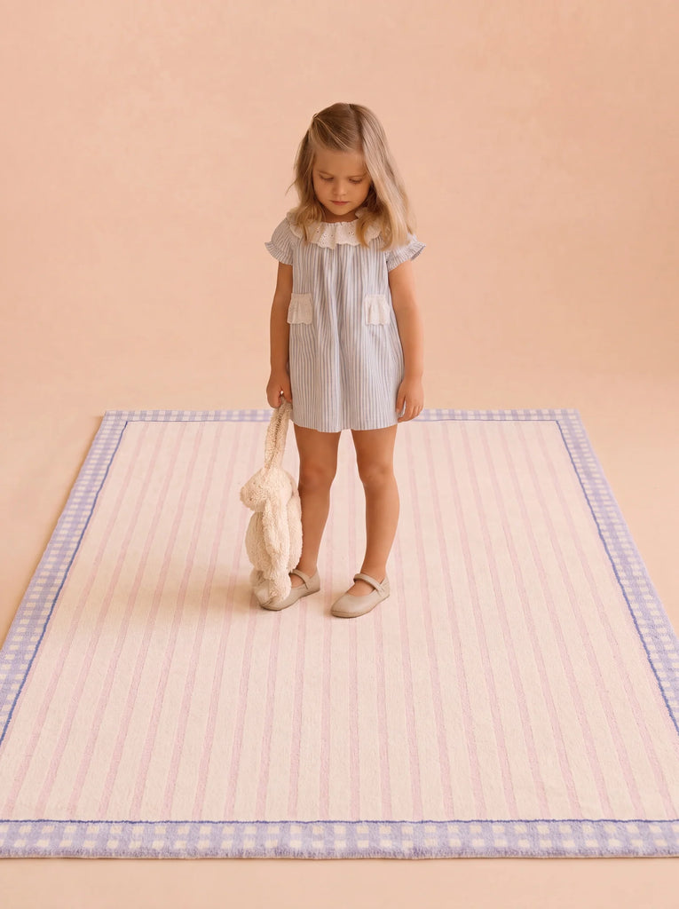 A young girl in a light blue dress stands on the Little Forest Animals Silk Rug — The Gingham, which features a charming blue gingham border, while holding a plush bunny. The background is plain beige.