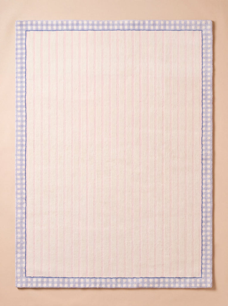 The "Little Forest Animals Silk Rug — The Gingham," a rectangular rug featuring pale pink vertical stripes and a blue gingham border evocative of hand-tufted detailing, is displayed flat on a neutral beige surface.