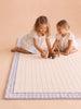 Two young girls sit on the Little Forest Animals Silk Rug — The Gingham, which features a pastel-striped design and a blue gingham border, playing with wooden animal toys. Both girls are smiling and wearing light-colored dresses in the softly lit space.
