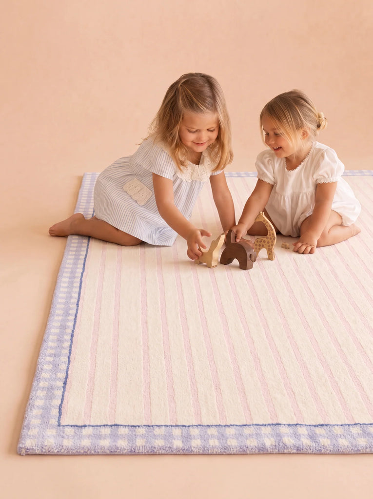 Two young girls sit on the Little Forest Animals Silk Rug — The Gingham, which features a pastel-striped design and a blue gingham border, playing with wooden animal toys. Both girls are smiling and wearing light-colored dresses in the softly lit space.