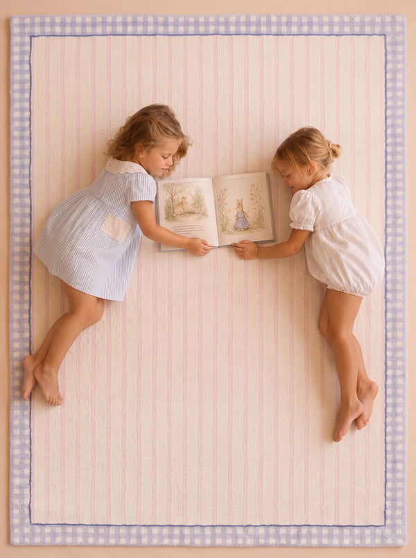 Two young girls in pastel dresses lie on the Little Forest Animals Silk Rug — The Gingham, with its soft hand-tufted texture and blue gingham border, facing each other as they read an illustrated book together.