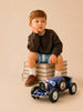 A young boy sits on a stack of books, resting his chin on his hand and looking thoughtful. He wears a dark sweater, blue shorts, white socks, and brown shoes. In front of him is a beautifully handcrafted die-cast car from the Little Forest Animals — The Grand Prix collection, capturing the timeless elegance of a Bugatti Type 35 model.