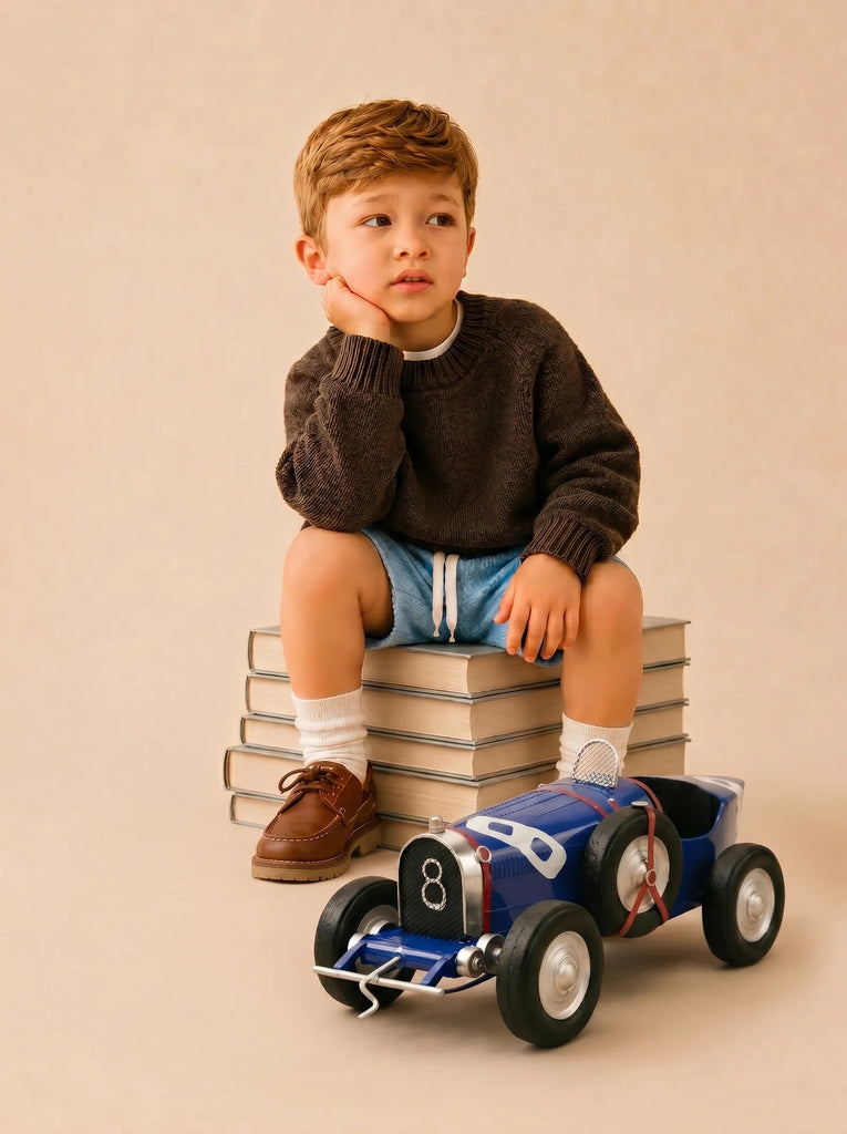 A young boy sits on a stack of books, resting his chin on his hand and looking thoughtful. He wears a dark sweater, blue shorts, white socks, and brown shoes. In front of him is a beautifully handcrafted die-cast car from the Little Forest Animals — The Grand Prix collection, capturing the timeless elegance of a Bugatti Type 35 model.