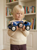 A young child with blonde hair holds "Little Forest Animals — The Grand Prix," a handcrafted die-cast car modeled after a blue Bugatti Type 35 with a propeller, standing in front of a bookshelf filled with colorful books.