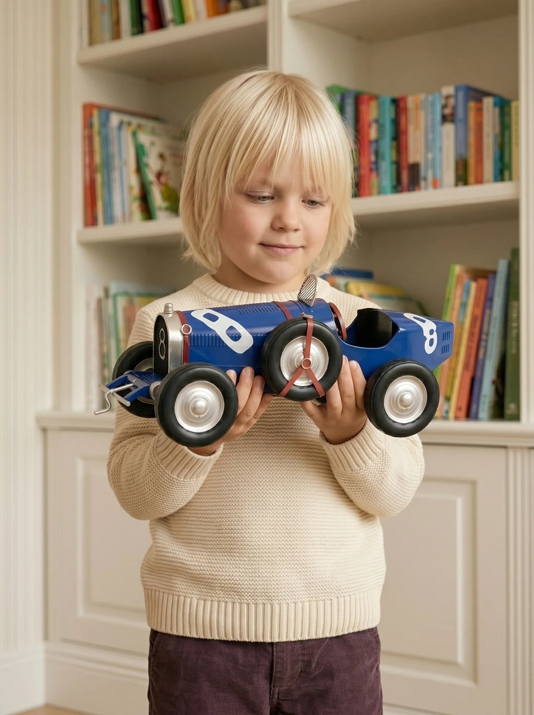 A young child with blonde hair holds "Little Forest Animals — The Grand Prix," a handcrafted die-cast car modeled after a blue Bugatti Type 35 with a propeller, standing in front of a bookshelf filled with colorful books.