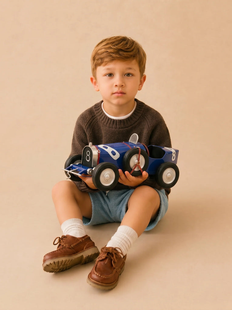 A young boy with light brown hair sits on the floor holding a Little Forest Animals — The Grand Prix handcrafted die-cast car. He wears a dark sweater, blue shorts, white socks, and brown shoes, gazing calmly at the camera against a plain beige background.