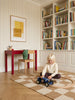 A young child sits on a checkered rug playing with Little Forest Animals — The Grand Prix in a cozy room featuring bookshelves, a red desk, a chair, and a framed abstract artwork on striped wallpaper.