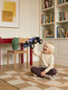 A young child sits on a checkered rug, smiling and holding up a Little Forest Animals — The Grand Prix handcrafted die-cast car. A wooden chair with a green cap, a red table, bookshelves, and a framed picture are in the background.