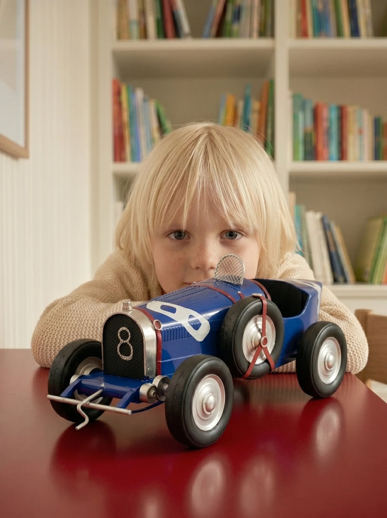 A young child with blond hair looks intently at Little Forest Animals — The Grand Prix, a handcrafted die-cast model car, on a red table, with bookshelves full of colorful books in the background.
