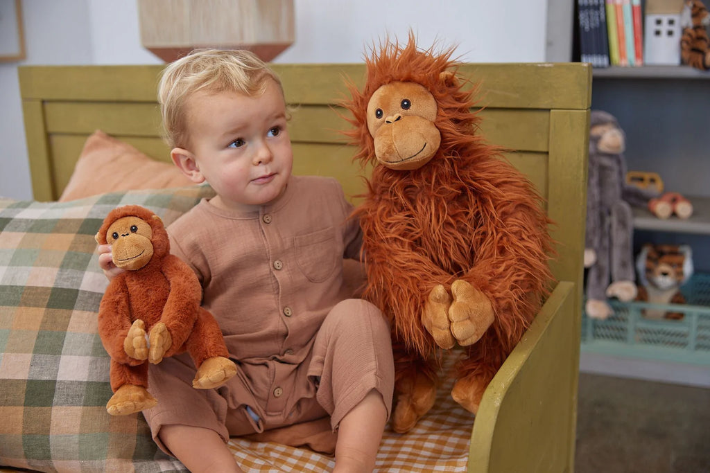 A young child in a brown outfit sits on a green bench holding two "The Orangutans Stuffed Animal" toys, one smaller and one larger, symbolizing a mother-daughter bond. The child gazes to the side with a thoughtful expression. In the background, there are stuffed animals and books on shelves.