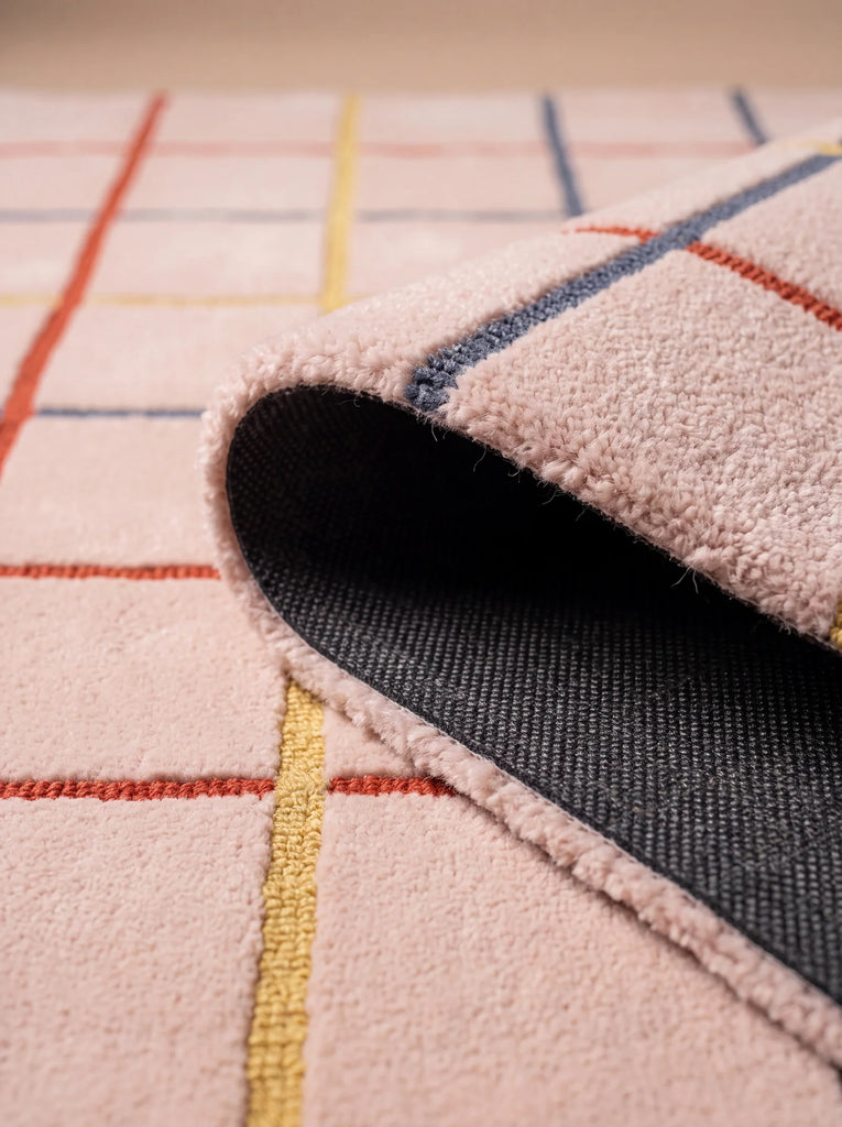 A close-up of the Little Forest Animals Silk Rug — The Palette showcases its vibrant design in light pink, accented with red, yellow, and blue grid lines. One corner is flipped to reveal a dark gray non-slip underside—perfect for adding comfort and safety to any designer kids room rug collection.