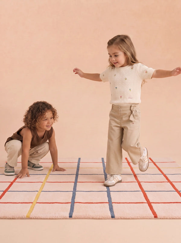 Two young children play on The Palette’s Little Forest Animals Silk Rug—a cream-colored, hand-tufted silk piece adorned with red, blue, and orange grid lines. One child stands with arms outstretched while the other crouches nearby, both dressed in cozy beige and brown attire—a perfect scene showcasing this stylish designer kids room rug.