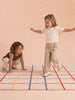 Two young children play on The Palette’s Little Forest Animals Silk Rug—a cream-colored, hand-tufted silk piece adorned with red, blue, and orange grid lines. One child stands with arms outstretched while the other crouches nearby, both dressed in cozy beige and brown attire—a perfect scene showcasing this stylish designer kids room rug.