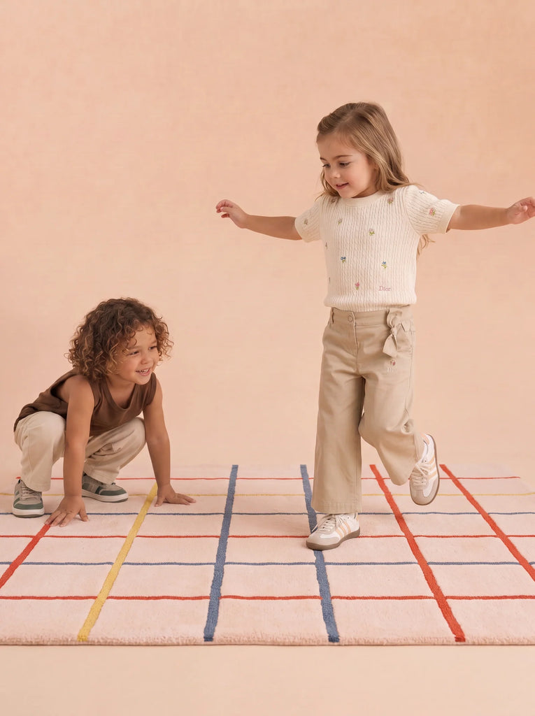 Two young children play on The Palette’s Little Forest Animals Silk Rug—a cream-colored, hand-tufted silk piece adorned with red, blue, and orange grid lines. One child stands with arms outstretched while the other crouches nearby, both dressed in cozy beige and brown attire—a perfect scene showcasing this stylish designer kids room rug.