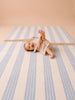 A smiling baby lies on their back on The Dachshund, a hand-tufted silk rug from the Little Forest Animals collection, holding one foot with their hand and looking up, dressed in a light outfit in a softly lit room with neutral tones.