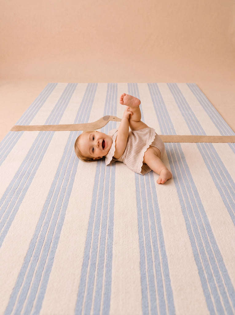 A smiling baby lies on their back on The Dachshund, a hand-tufted silk rug from the Little Forest Animals collection, holding one foot with their hand and looking up, dressed in a light outfit in a softly lit room with neutral tones.