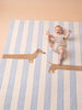 A smiling baby in a light romper lies on the Little Forest Animals Silk Rug — The Dachshund, a striped children’s room rug featuring playful dachshund illustrations, with one design cleverly positioned so the baby’s legs appear as the dog’s legs.