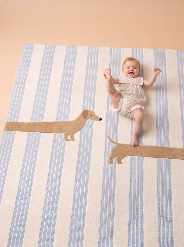 A smiling baby in a light romper lies on the Little Forest Animals Silk Rug — The Dachshund, a striped children’s room rug featuring playful dachshund illustrations, with one design cleverly positioned so the baby’s legs appear as the dog’s legs.
