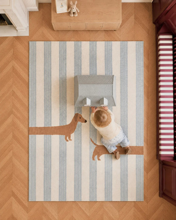 A baby sits on the Little Forest Animals Silk Rug — The Dachshund, playing with a toy house. The room features a wood floor and built-in seating with a red-and-white striped cushion, making this rug the perfect cozy addition for children’s playtime.
